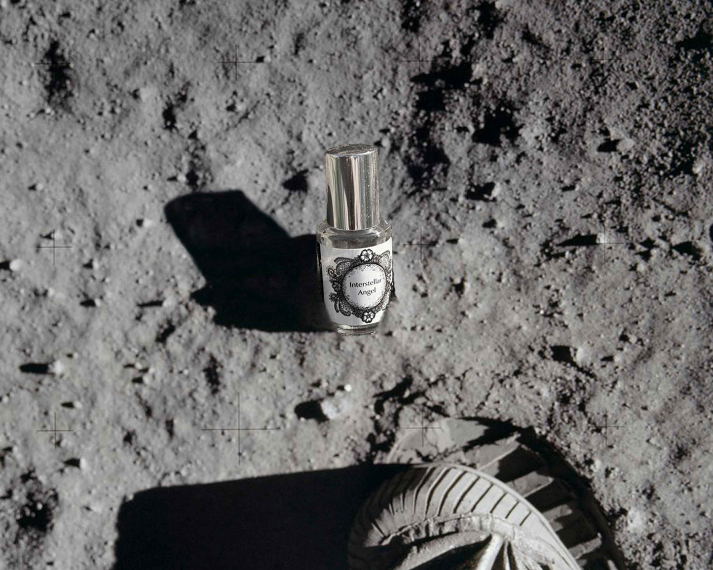 5 mL glass perfume bottle with silver cap of the scent "Interstellar Angel" half-buried in moon dust in front of the toe of an astronaut boot.