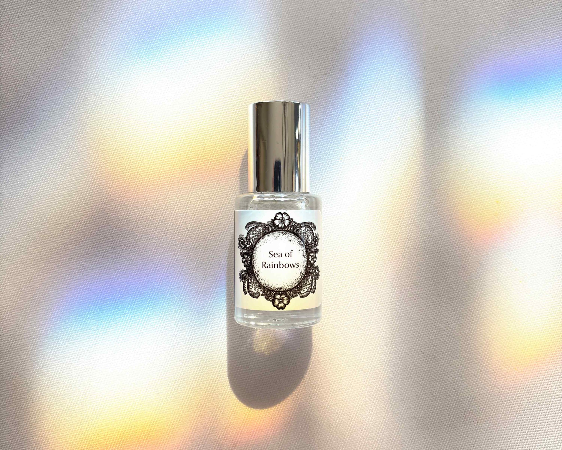 5 mL glass perfume bottle with silver cap of the scent "Sea of Rainbows" on a white cloth with soft prismatic rainbows.
