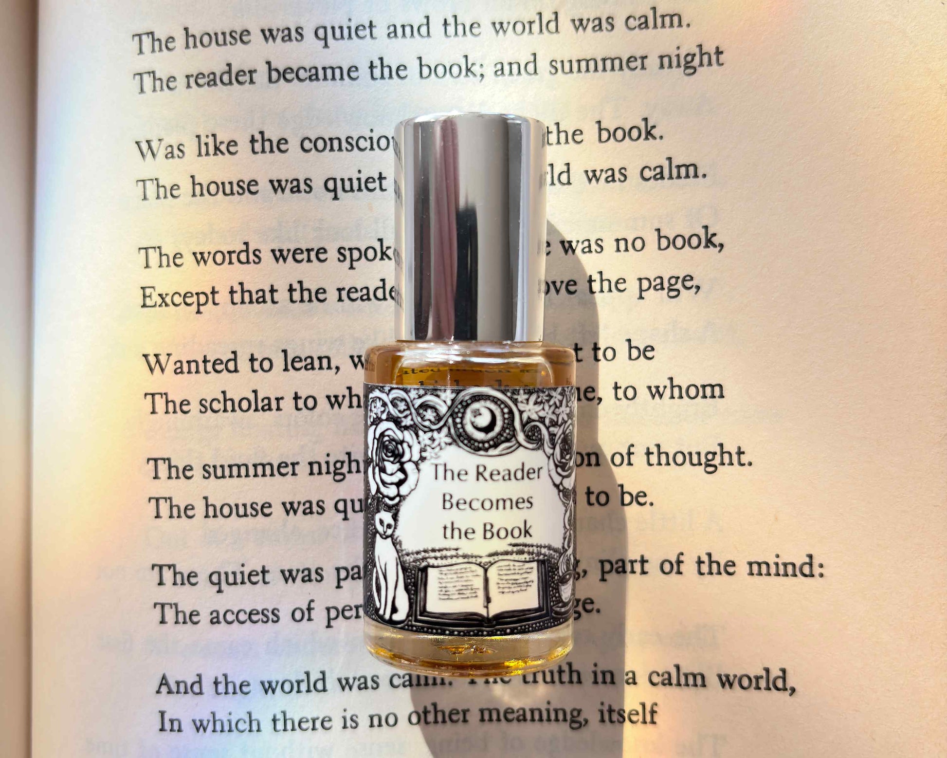 5 mL glass perfume bottle with silver cap of the scent "The Reader Becomes the Book" laid atop a book open to the poem it was inspired by.